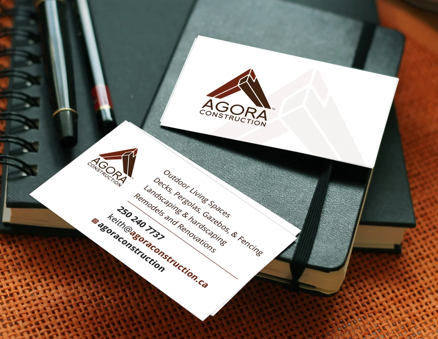 A stack of Agora Construction business cards on a dark leather Moleskine notebook, the top card showing the full cedar-and-brown A-frame mark with AGORA CONSTRUCTION beneath, the lower card showing contact details for Keith Grant at agoraconstruction.ca with service categories including Outdoor Living Spaces, Decks, Pergolas, Gazebos, Fencing, Landscaping, and Remodels